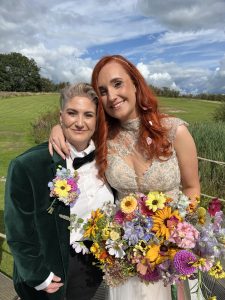 A couple with arms around each other. One has dress and flowers and the other a fab evening suit.